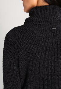 Suéter negro de punto con un cuello alto y un patrón texturizado, con un pequeño logo de la marca en la parte posterior.