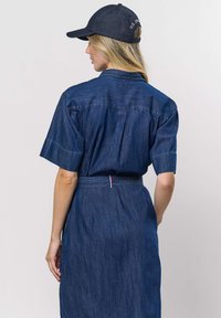 Femme aux longs cheveux blonds portant une robe en jean bleu foncé et une casquette de baseball bleu marine, vue de dos sur un fond uni.