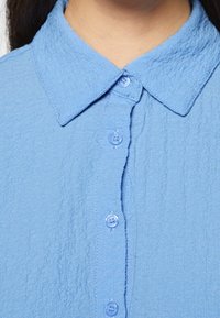 Chemise bleu clair, texturée, avec un col classique et cinq boutons assortis sur le devant. Le tissu semble légèrement froissé.