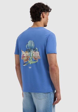 Man met zijn rug naar de camera, draagt een blauw T-shirt met bloemmotief en tekst "Pure Path" op de rug, gecombineerd met lichtblauwe spijkerbroek.
