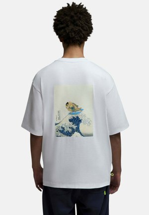 Personne portant un T-shirt blanc oversize avec une vague bleue et un graphisme de lutteur sumo surfant dans le dos, debout dos à la caméra.