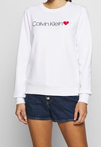 Felpa bianca con maniche lunghe e polsini a coste, che presenta il logo "Calvin Klein" in nero e una grafica a forma di cuore in rosso.