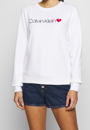 Sudadera blanca de manga larga con puños de punto, que presenta el logo de "Calvin Klein" en negro y un gráfico de corazón en rojo.