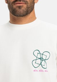 Biely sweatshirtn so zelenou grafikou dronu a ružovým vlnovým logom. Tkanina vyzerá mäkko a štruktúrovane s okrúhlym výstrihom.