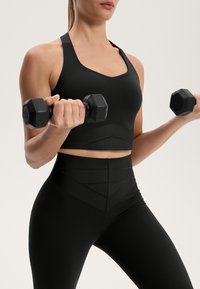 Črn športni crop top in visoko pasne legice z teksturiranimi detajli. Model drži dva šestkotna črna dumbbell.