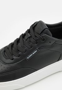 Czarne skórzane sneakersy z teksturowaną powierzchnią, płaskimi czarnymi sznurowadłami, perforacjami w pobliżu palców oraz białą gumową podeszwą. Zawierają etykietę marki.