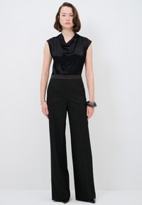 Haut en satin noir avec un col drapé, associé à un pantalon noir à taille haute et jambes larges. Bracelet jonc transparent au poignet.