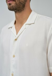 Camicetta bianca a maniche corte realizzata in tessuto leggero. Caratterizzata da una chiusura con bottoni, un logo discreto sul petto e un design classico del colletto.