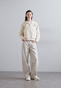 Casaco bomber cropped bege com fecho metálico e bolsos frontais, combinado com calças largas cinza-claro e ténis brancos.