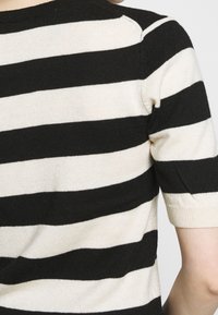 Osoba oblečená v černobíle pruhovaném pleteném tričku s krátkým rukávem, zobrazená ze zadní strany a oblasti ramene.