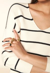 Pull rayé crème et marine avec un col en V. Mains ornées de trois bagues en or, présentant diverses formes et textures.