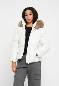 Vrouw in een witte pufferjack met een met bont afgewerkte capuchon, zwarte top en grijze cargobroek, staande met één hand in de zak.