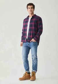 Chemise en flanelle à carreaux bleu marine et bordeaux, portée sur un t-shirt blanc, associée à un jean bleu clair et des bottes de travail beige.
