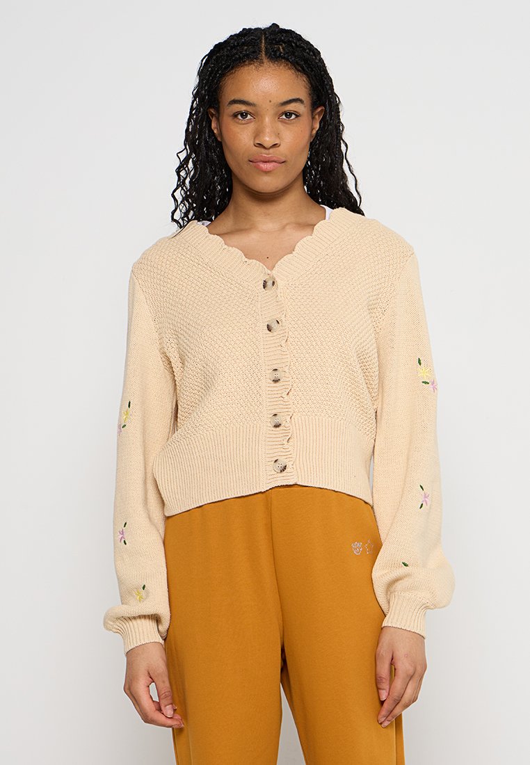 Beige gebreide cardigan met gegolfde halslijn en knoopsluiting; heeft bloemenborduurwerk op de mouwen; cropped ontwerp met ribbelboord.