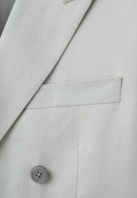 Gros plan sur le tissu d'un blazer gris clair montrant un revers à encoche, une poche passepoilée et un bouton gris à quatre trous sur le devant.