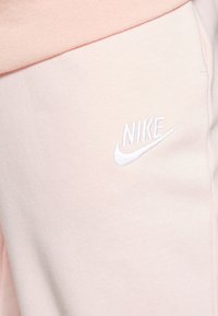 Šviesiai rožiniai Nike treningai su subtilia tekstūra, elastinga juosta ir baltu siuvinėjimu, esančiu šalia kišenės.