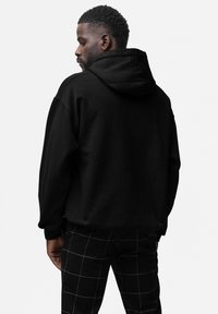 Uomo con capelli ricci corti, che indossa un hoodie nero e pantaloni neri con linee bianche a griglia, in piedi di lato su uno sfondo bianco.