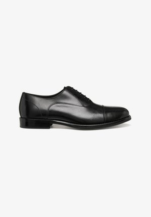 Zapato de vestir Oxford de cuero negro con cordones cerrados, puntera con tapa, tacón bajo y acabado liso y pulido, mostrado en perfil lateral sobre fondo blanco.