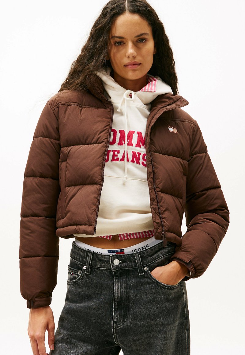 Giacca puffer corta marrone con un design trapuntato. Presenta una chiusura a zip frontale, collo alto ed è indossata sopra un hoodie bianco con logo rosso.