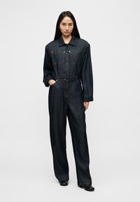 Combinaison en denim bleu foncé, avec une fermeture à boutons sur le devant, un col, des manches longues et deux poches poitrine, offrant une coupe décontractée.