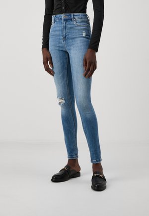 Skinny jeans in light blue denim with a high waist, featuring distressed details and a slight fade. Paired with black loafers.