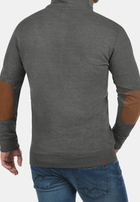 Felpa grigia con collo alto, caratterizzata da toppe marroni sui gomiti e polsini a coste. Texture morbida, design aderente e orlo di lunghezza media.