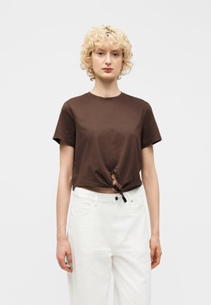 Osoba o blond, kręconych włosach, ubrana w brązowy krótki top z węzłem oraz białe, wysokie spodnie, stoi na tle gładkiego tła.