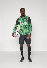 Grøn og sort langærmet fodboldtrøje med camouflage-mønster, sorte shorts og sorte sokker. Spilleren holder en fodbold.