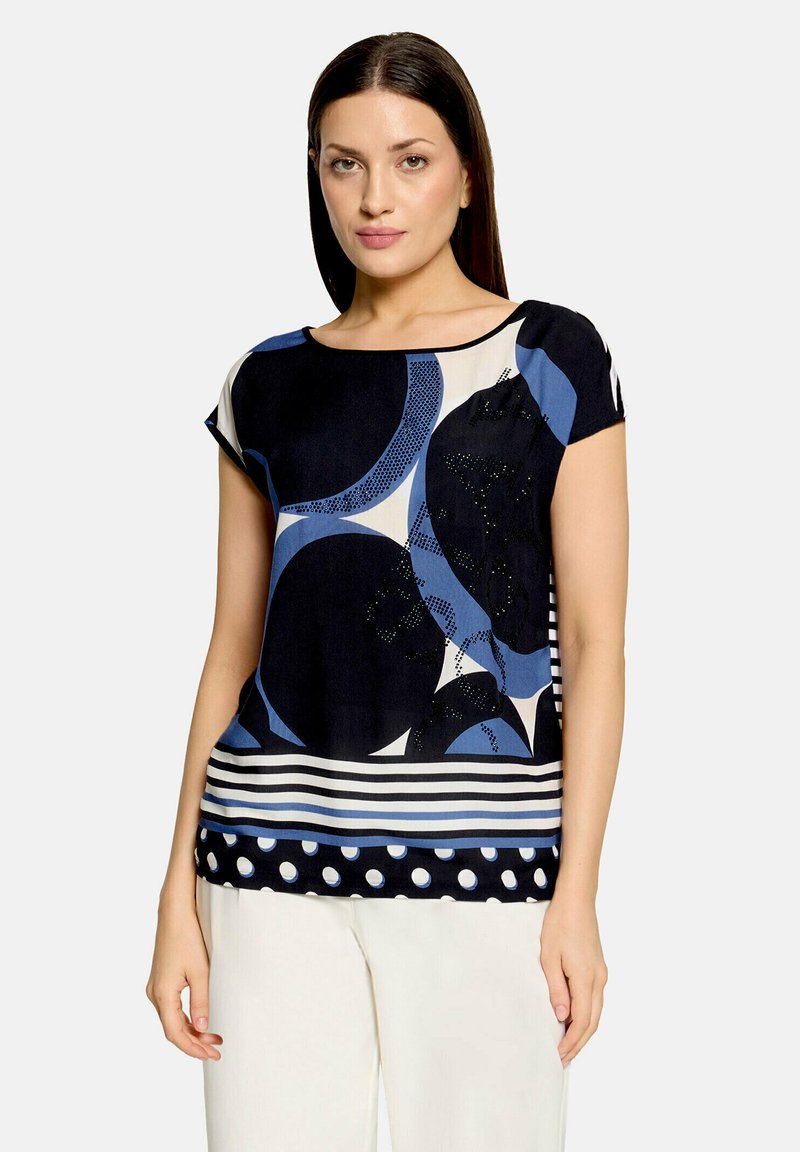 Femme portant un haut bleu marine à manches courtes avec des formes abstraites bleues et crème, des rayures et des pois, assorti à un pantalon blanc.