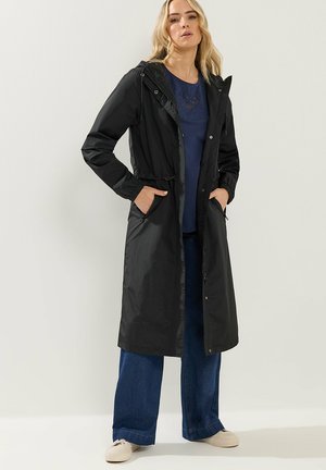 Kobieta w długim czarnym płaszczu przeciwdeszczowym z kapturem, nałożonym na granatową koszulę i szerokie niebieskie dżinsy, z rękami w kieszeniach płaszcza na gładkim tle.