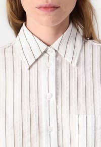 Camicia bianca con bottoni, a righe nere sottili, con colletto ampio, patta frontale e una singola tasca sul petto. Tessuto testurizzato.