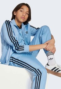 Világoskék Adidas melegítőfutóruha fekete csíkokkal, cipzáras dzsekivel és hozzáillő nadrággal, fehér cipővel és csíkos zoknival viselve.