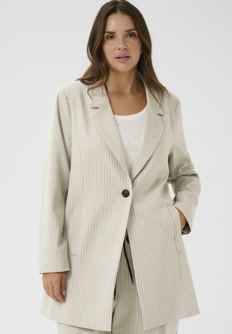 Mujer con una chaqueta de rayas beige con un botón abrochado sobre una blusa blanca, de pie contra un fondo claro y uniforme.