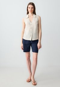 Camicetta beige senza maniche in lino abbinata a pantaloni corti blu scuro. Design semplice, texture leggera e vestibilità rilassata creano un look informale.