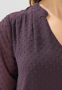 Fioletowa, teksturowana bluzka z małymi, wypukłymi kropkami. Dekolt w serek z zaokrąglonym kołnierzykiem, przejrzysty materiał z lekko połyskującą powierzchnią oraz subtelne fałdy.