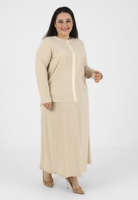 Abito beige a maniche lunghe con dettagli a bottoni testurizzati lungo la parte anteriore, realizzato in morbido tessuto con una silhouette fluida. Indossato con tacchi nudi.