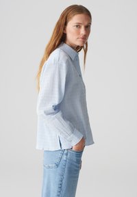 Camicetta a righe azzurre con colletto button-down, maniche lunghe e vestibilità rilassata. Il tessuto ha una finitura testurizzata. Indossata con jeans blu.
