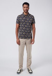 Camicia a maniche corte di colore blu navy con un motivo floreale giallo e bianco, abbinata a pantaloni beige chiaro e sneaker bianco e nero.