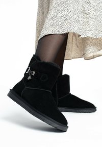 Botas de tobillo de ante negro con un forro interior suave, que presentan un charm plateado decorativo y una suela de goma baja. Adecuadas para el uso casual.