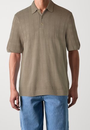 Mężczyzna w oliwkowej koszulce polo z krótkim rękawem i fakturowanymi kwadratami oraz jasnoniebieskich jeansach, stojący na tle jednolitego tła.