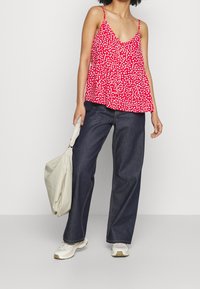 Donna che indossa un top rosso senza maniche con pois bianchi, jeans a gamba larga blu scuro, sneakers bianche e tiene una borsa beige.