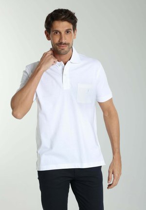 Homme aux cheveux courts et foncés, portant un polo blanc et un pantalon foncé, touchant son cou, debout devant un fond clair uni.