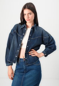 Džinsinis švarkas su sagų uždarymu, ilgomis rankovėmis ir laisvu siluetu. Tamsiai mėlyna spalva su kontrastingais siūlais ir apykakle.