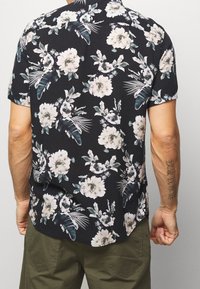 Camisa negra de manga corta con un patrón floral que presenta flores de color claro y hojas verdes; un ajuste holgado y casual con un dobladillo redondeado.