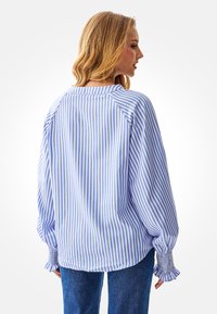 Blauw-wit gestreepte blouse met ballonmouwen en gerimpelde manchetten. Heeft een ronde halslijn en licht geweven stof.