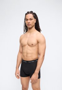 Άνδρας με μακριές κοτσίδες, φορώντας μαύρα μποξεράκια Calvin Klein, στέκεται κορμός γυμνός μπροστά σε απλό φωτεινό φόντο.