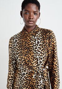 Abito camicia con stampa leopardata nei colori nero, arancione e crema. Presenta un colletto, bottoni e una cintura da annodare, realizzato in un tessuto liscio.