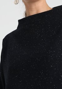 Svart, texturerad topp med hög halsringning, prydd med ett subtilt glittermönster. Slätt tyg med en lös och avslappnad passform.