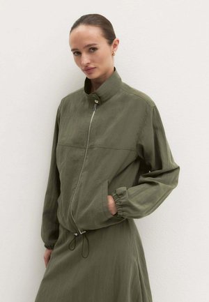 Donna con i capelli raccolti che indossa una giacca verde oliva ampia con zip e gonna abbinata su sfondo chiaro.