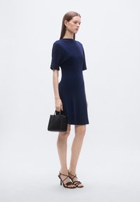 Rochie albastru marin cu mâneci scurte, croială lejeră, asortată cu pantofi cu toc înalt negri, cu barete, și o geantă mică neagră. Textură netedă a materialului.